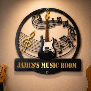 Könnte beinhalten: Ein rundes Wandbild aus schwarzem Metall mit einer schwarzen E-Gitarre in der Mitte, einem goldenen Violinschlüssel und schwarzen Noten auf Notenlinien. Ein schwarzes rechteckiges Schild unten zeigt den goldenen Text JAMES'S MUSIC ROOM.