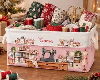 Puede incluir: Una cesta de almacenamiento rectangular de tela con asas de cuerda, con una base rosa con una escena de taller de costura impresa y el nombre Emma. La cesta está llena de artículos de costura, incluyendo rollos de tela, hilos y tijeras.
