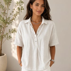 May include: A short-sleeved, off-white button-down shirt with a collar and a relaxed fit. The shirt features a partial button placket down the center front, light-colored buttons, and a textured fabric finish. The design includes a simple hemline.