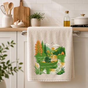 May include: A beige kitchen towel featuring a colorful embroidered graphic of a person in a green hat sitting in a green canoe on a lake, fishing with a rod. The scene includes orange and green trees, mountains, a yellow sun, and cattails.
