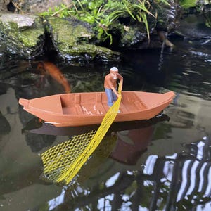 Pode incluir: Um barco miniatura castanho a flutuar na água, contendo uma pequena figura com um chapéu branco, camisa castanha e calças azuis. A figura segura uma rede de pesca de malha amarela que se estende para a água a partir do barco.