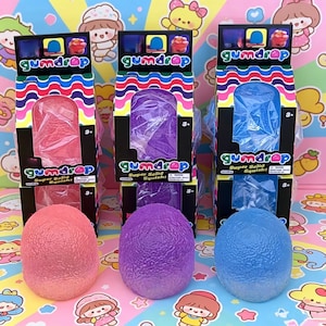 May include: Three Gumdrop Super Solid Squish toys in pink, purple, and blue, each packaged in a black box with a colorful wavy pattern at the top. The boxes feature the text Gumdrop and Super Solid Squish, with clear plastic windows.