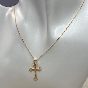 May include: A gold-toned metal cross pendant suspended from a matching delicate gold-toned chain. The cross features a decorative, ornate design with flared ends and a textured, twisted rope-like detail running vertically through the center.