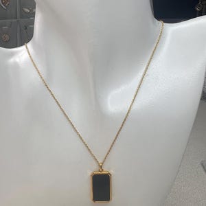 May include: A gold-toned necklace featuring a thin, twisted chain and a rectangular pendant. The pendant has a polished gold-toned frame surrounding a smooth, solid black rectangular center piece, suspended from a small gold-toned bail.