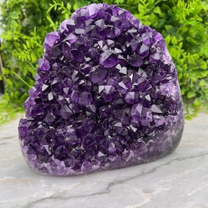 Pode incluir: Uma grande geodo de ametista com pontas de cristal facetadas de cor púrpura profunda agrupadas. A estrutura tem uma forma arredondada e irregular com uma base sólida e lisa, exibindo um tom violeta vibrante e saturado em todo o conjunto.