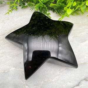 Peut inclure: Un objet en forme d'étoile polie avec une surface noire lisse et réfléchissante. L'étoile à cinq branches présente des bords nets et précis ainsi qu'une finition brillante qui crée un effet miroir, reflétant l'environnement sur sa surface sombre.