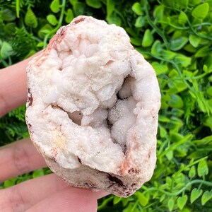 Pode incluir: Uma geoda de quartzo rosa pálido bruto com uma cavidade central preenchida por pequenas estruturas cristalinas cintilantes. O exterior da pedra apresenta uma textura áspera e irregular com depósitos minerais castanho-avermelhados.