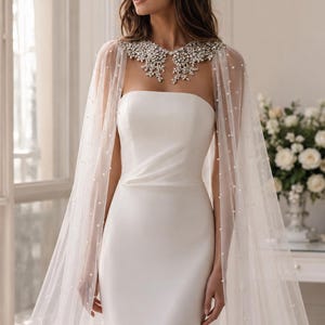 May include: A floor-length white strapless gown featuring a smooth, fitted bodice and a sheer, floor-length cape overlay. The cape is adorned with scattered white pearls and features a decorative collar embellished with clear crystals and beads.