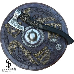 May include: A round wooden shield with a dark brown finish, featuring intricate gold-colored knotwork, a central silver-toned metal boss, and engraved runes along the rim. A matching axe with a silver blade and dark handle rests on top.