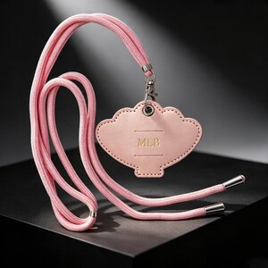 May include: A light pink shell-shaped badge holder with the letters MLB embossed in gold. The holder is attached to a matching light pink cord lanyard featuring silver-toned metal hardware at the clasp and the ends of the cord.