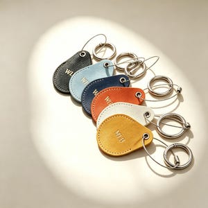 May include: A collection of six teardrop-shaped leather keychains in black, light blue, navy, orange, white, and yellow. Each keychain features a metal grommet, a thin wire cable, and a circular metal ring. The leather is embossed with the letters MLB.