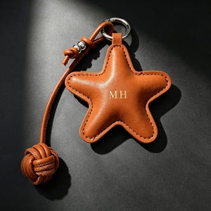 May include: A star-shaped leather keychain in a warm tan color featuring the embossed letters MH in the center. The keychain includes a silver-toned metal ring, a long leather cord with a knotted end, and small silver-toned metal beads.