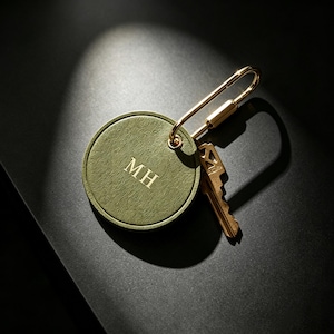 Pode incluir: Um porta-chaves redondo de cor verde-azeitona com textura e as letras MH gravadas no centro. A etiqueta está presa a um mosquetão de metal dourado brilhante que segura uma chave de cor latão, criando um acessório funcional.