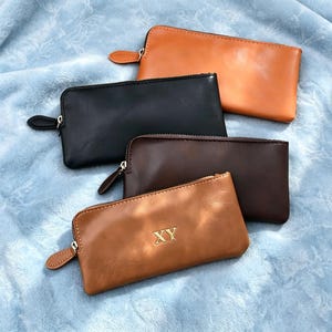 May include: Four rectangular zippered pouches in tan, black, dark brown, and a lighter tan. The lighter tan pouch features the gold-colored letters XY embossed in the center. Each pouch has a matching zipper pull tab and a smooth finish.