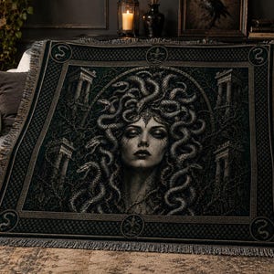May include: A woven throw blanket featuring a dark, detailed illustration of Medusa with snakes for hair. The design includes architectural stone pillars in the background, a decorative border with small snake motifs, and a fringed edge finish.