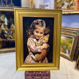 Könnte beinhalten: Ein rechteckiges gerahmtes Gemälde, das ein junges Kind mit hellbraunem Haar in einem gemusterten Kleid zeigt, das ein flauschiges orangefarbenes getigertes Kätzchen hält. Das Kunstwerk befindet sich in einem dicken, verzierten Goldrahmen.