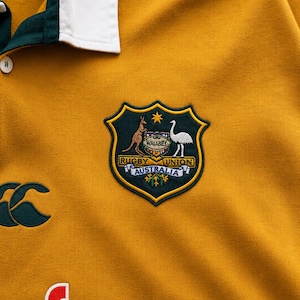 May include: A mustard yellow rugby shirt with a white collar and dark green trim. It features an embroidered Wallabies Rugby Union Australia crest, a dark green Canterbury logo, and the partial red and white text Vodafone printed on the front.