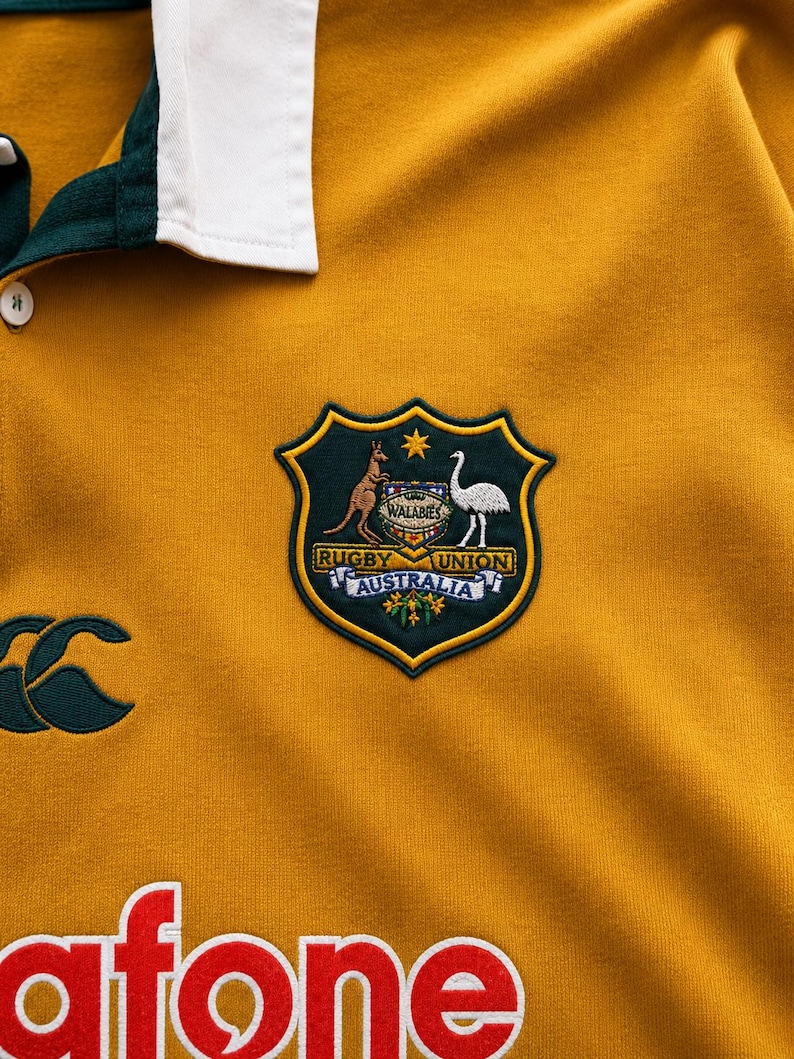 May include: A mustard yellow rugby shirt with a white collar and dark green trim. It features an embroidered Wallabies Rugby Union Australia crest, a dark green Canterbury logo, and the partial red and white text Vodafone printed on the front.