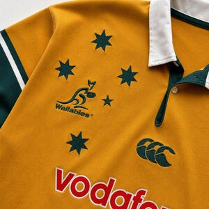 May include: A gold rugby shirt with a white collar and dark green sleeves. The front features the Wallabies logo, a kangaroo graphic, five dark green stars, the Canterbury logo, and the word Vodafone in red letters with a white outline.