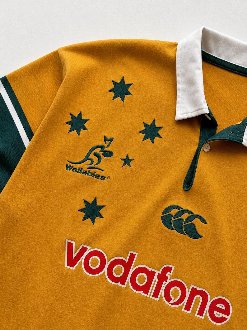 May include: A gold rugby shirt with a white collar and dark green sleeves. The front features the Wallabies logo, a kangaroo graphic, five dark green stars, the Canterbury logo, and the word Vodafone in red letters with a white outline.