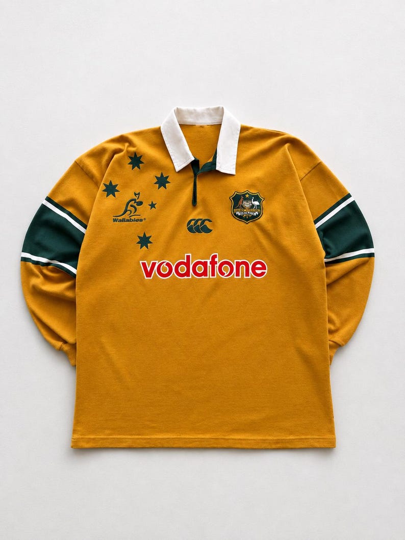 May include: A gold long-sleeve rugby shirt with a white collar, green sleeve bands, and the text "vodafone" in red. It features the Wallabies logo, the Australian coat of arms, and the Southern Cross constellation in dark green on the chest.