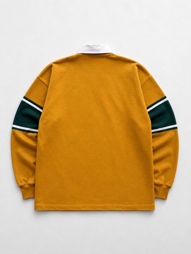 May include: A long-sleeve rugby shirt in mustard yellow featuring a white collar and horizontal dark green bands on the sleeves, accented by thin white stripes bordering the green sections, with ribbed cuffs and a straight hemline.