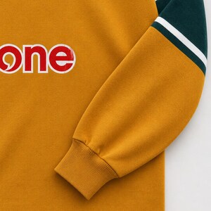 May include: A mustard yellow long-sleeve sweatshirt featuring a dark green and white striped sleeve detail, a ribbed cuff, and a partial graphic print of the word one in white letters. A circular Australia emblem is visible at the neckline.