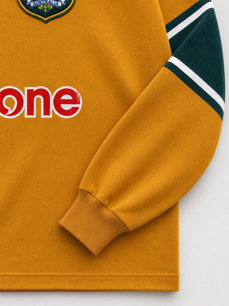 May include: A mustard yellow long-sleeve sweatshirt featuring a dark green and white striped sleeve detail, a ribbed cuff, and a partial graphic print of the word one in white letters. A circular Australia emblem is visible at the neckline.