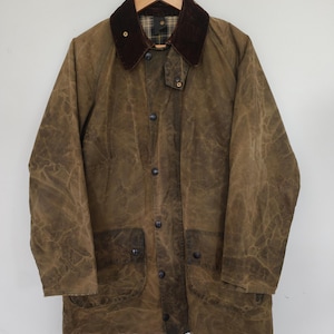 May include: A brown waxed cotton jacket featuring a dark brown corduroy collar, a front button placket with metal snap closures, and two large front patch pockets. The interior lining displays a classic plaid pattern in shades of brown and green.