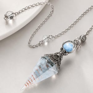 Könnte beinhalten: Ein Pendel an einer silberfarbenen Metallkette mit einem facettierten, kegelförmigen, klaren Stein, der innen Kupferdrahtspiralen enthält. Die Spitze des Steins ist mit verziertem silbernem Metall und einer kleinen, runden, hellblauen Perle versehen.