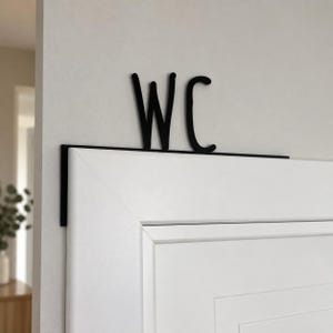 Könnte beinhalten: Ein Türschild aus schwarzem Metall mit den Buchstaben WC in einer stilisierten Schriftart. Das Schild hat eine flache, horizontale Basis, die über die Oberkante einer weißen Tür greift, sodass die Buchstaben über dem Türrahmen aufrecht stehen.