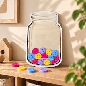 Könnte beinhalten: Ein gläserförmiges Holzdisplay mit einer transparenten Vorderseite, das mehrere kleine, bunte, runde Spielsteine mit eingeprägten Smileys enthält. Weitere lose Smiley-Spielsteine in Pink, Gelb, Blau und Lila liegen unterhalb des Displays.