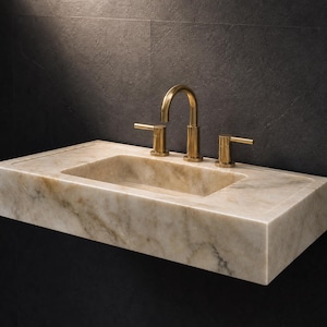 May include: A rectangular wall-mounted bathroom sink crafted from beige and cream-colored natural stone with visible veining. The sink features a central basin and is equipped with a three-piece faucet set finished in a brushed metallic gold tone.