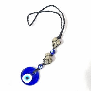 Peut inclure: Une décoration suspendue composée d'un cordon noir, de deux pierres de pyrite métalliques dans un filet noir et de deux perles en verre bleu en forme d'œil. La perle œil circulaire plus grande est en bas, avec une perle plus petite au milieu.
