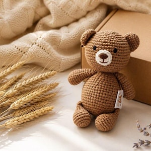 May include: A small, brown, crocheted teddy bear with a round head, two ears, and a white muzzle featuring a dark brown stitched nose and mouth. The bear has black bead eyes and a small white fabric tag attached to its side with a bear illustration.