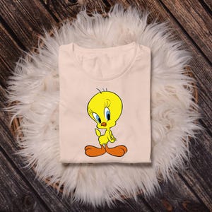 May include: A light peach-colored t-shirt featuring a graphic print of the yellow cartoon character Tweety Bird. The character has large blue eyes, orange feet, and a small tuft of hair on its head, centered on the front of the garment.
