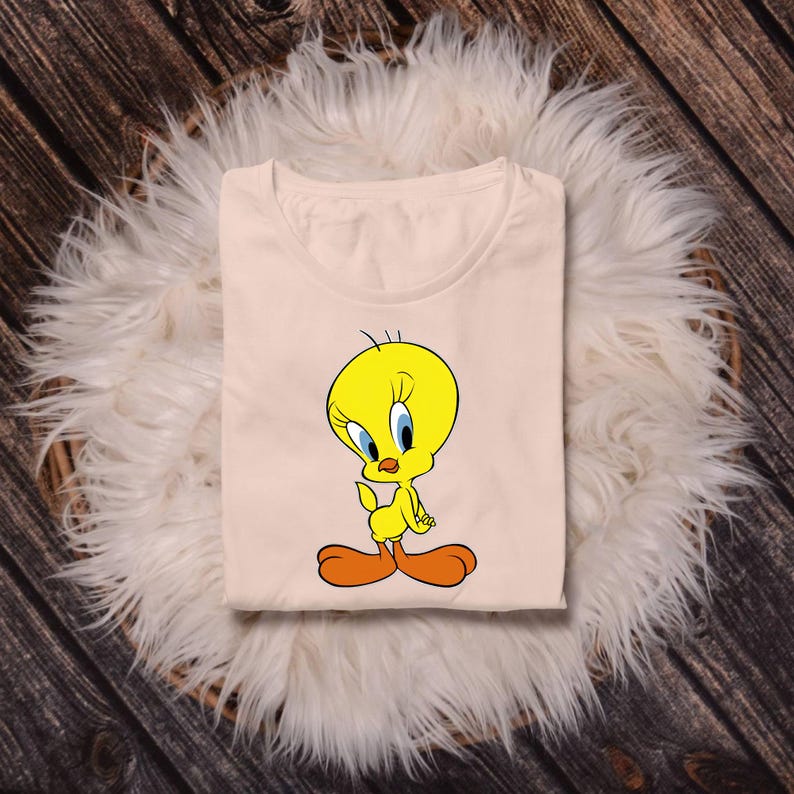 May include: A light peach-colored t-shirt featuring a graphic print of the yellow cartoon character Tweety Bird. The character has large blue eyes, orange feet, and a small tuft of hair on its head, centered on the front of the garment.