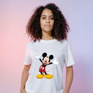 May include: A white short-sleeved t-shirt featuring a graphic print of Mickey Mouse on the front. The character is depicted with black ears, a flesh-toned face, red shorts with two white buttons, white gloves, and large yellow shoes, standing with arms outstretched.