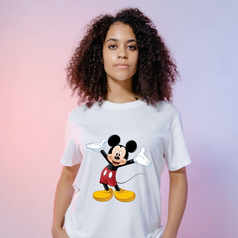 May include: A white short-sleeved t-shirt featuring a graphic print of Mickey Mouse on the front. The character is depicted with black ears, a flesh-toned face, red shorts with two white buttons, white gloves, and large yellow shoes, standing with arms outstretched.