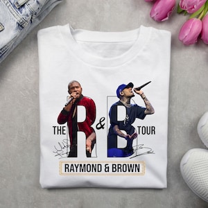 May include: A white t-shirt featuring a graphic print of two singers performing with microphones. The design includes the text THE R&B TOUR and RAYMOND & BROWN, along with two stylized signatures. The singers are wearing casual clothing.
