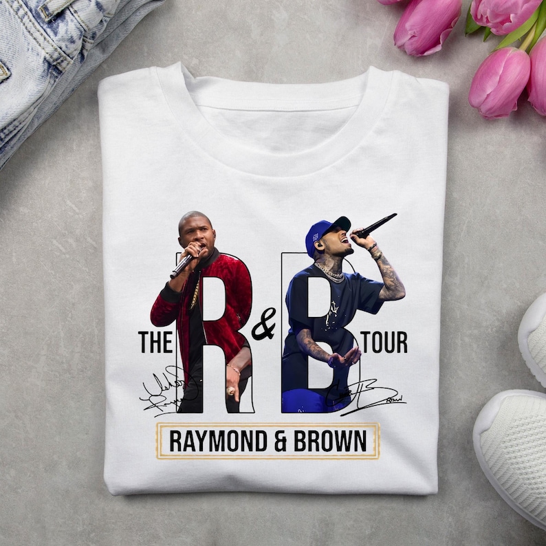 May include: A white t-shirt featuring a graphic print of two singers performing with microphones. The design includes the text THE R&B TOUR and RAYMOND & BROWN, along with two stylized signatures. The singers are wearing casual clothing.