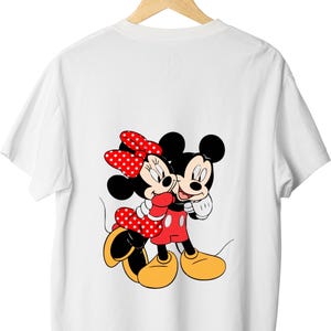 May include: A white short-sleeved t-shirt featuring a graphic print of Mickey Mouse and Minnie Mouse embracing. Minnie wears a red dress with white polka dots and a matching bow, while Mickey wears red shorts and yellow shoes.