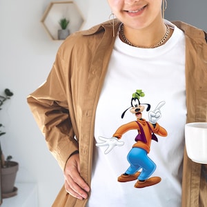 May include: A white t-shirt featuring a graphic print of the cartoon character Goofy, who is wearing an orange long-sleeve shirt, a purple vest, blue pants, and brown shoes, while pointing upward with one white-gloved hand.