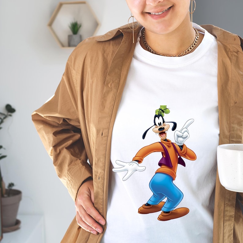 May include: A white t-shirt featuring a graphic print of the cartoon character Goofy, who is wearing an orange long-sleeve shirt, a purple vest, blue pants, and brown shoes, while pointing upward with one white-gloved hand.
