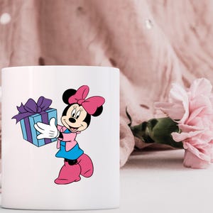 May include: A white ceramic mug featuring a graphic of Minnie Mouse wearing a pink bow, a pink jacket, and a blue skirt, holding a light blue gift box with a purple ribbon. The character has white gloves and pink shoes, depicted in a classic style.