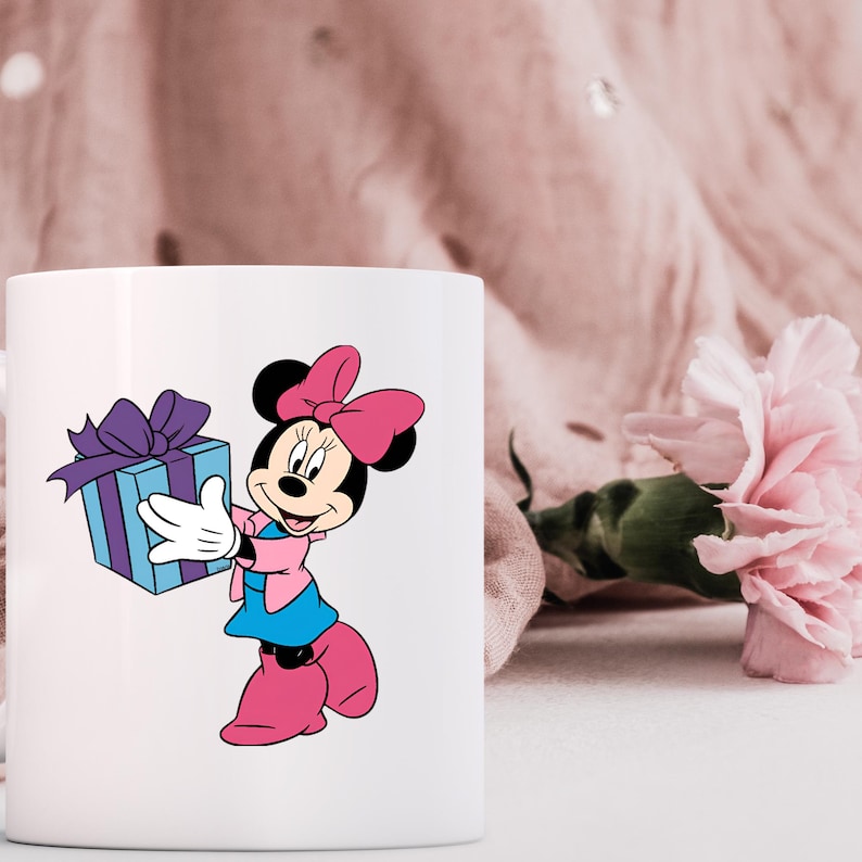 May include: A white ceramic mug featuring a graphic of Minnie Mouse wearing a pink bow, a pink jacket, and a blue skirt, holding a light blue gift box with a purple ribbon. The character has white gloves and pink shoes, depicted in a classic style.