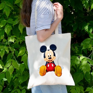 May include: A cream-colored canvas tote bag featuring a centered, full-color graphic of Mickey Mouse sitting down. The character is depicted with his signature black ears, red shorts with two white buttons, and large yellow shoes on a plain background.