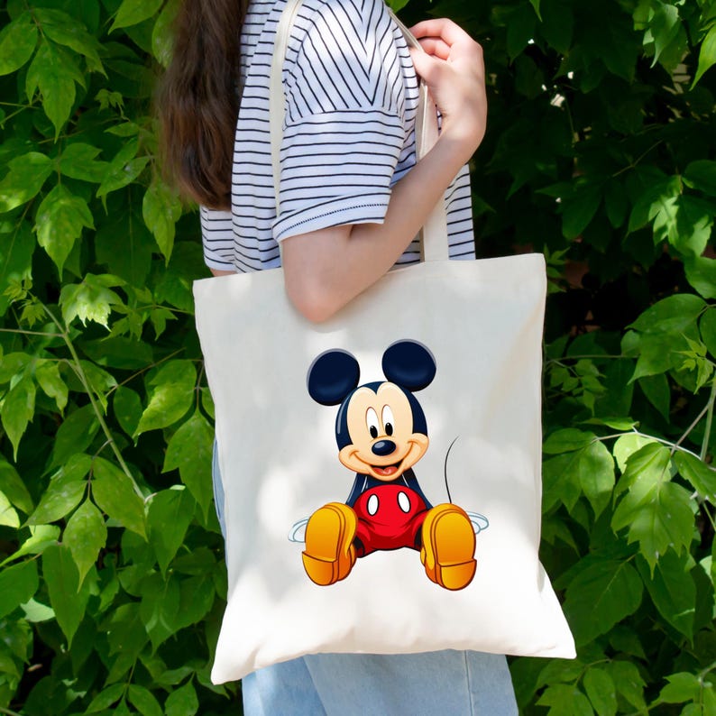 May include: A cream-colored canvas tote bag featuring a centered, full-color graphic of Mickey Mouse sitting down. The character is depicted with his signature black ears, red shorts with two white buttons, and large yellow shoes on a plain background.
