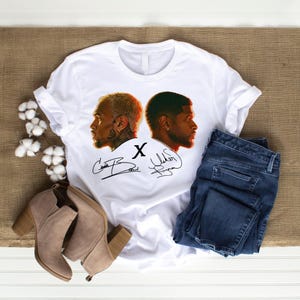 May include: A white t-shirt featuring a graphic of two men in profile facing away from each other, with the letter X in the center and two black signatures below. The shirt is displayed alongside a pair of dark blue denim jeans and tan boots.