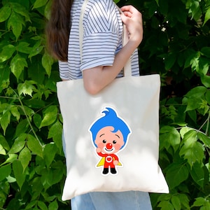 May include: A cream-colored canvas tote bag featuring a graphic print of a cartoon character with bright blue hair, a red nose, a red shirt with a white heart emblem, black pants, and a yellow cape, positioned centrally on the front of the bag.